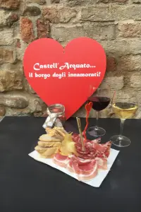 Tagliere di salumi piacentini, gnocco fritto e calici di vino sotto un cuore rosso con la scritta “Castell’Arquato… il borgo degli innamorati!”